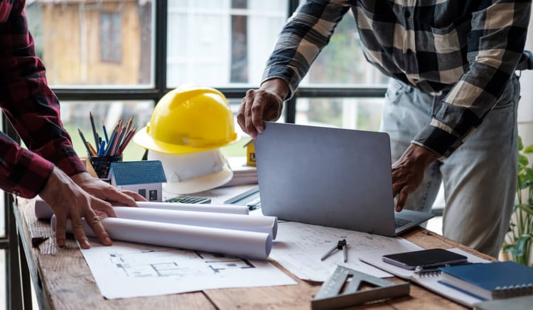 Construction professionals strategize using HubSpot CRM on a laptop amidst blueprints and tools on a worktable, highlighting digital management in construction.