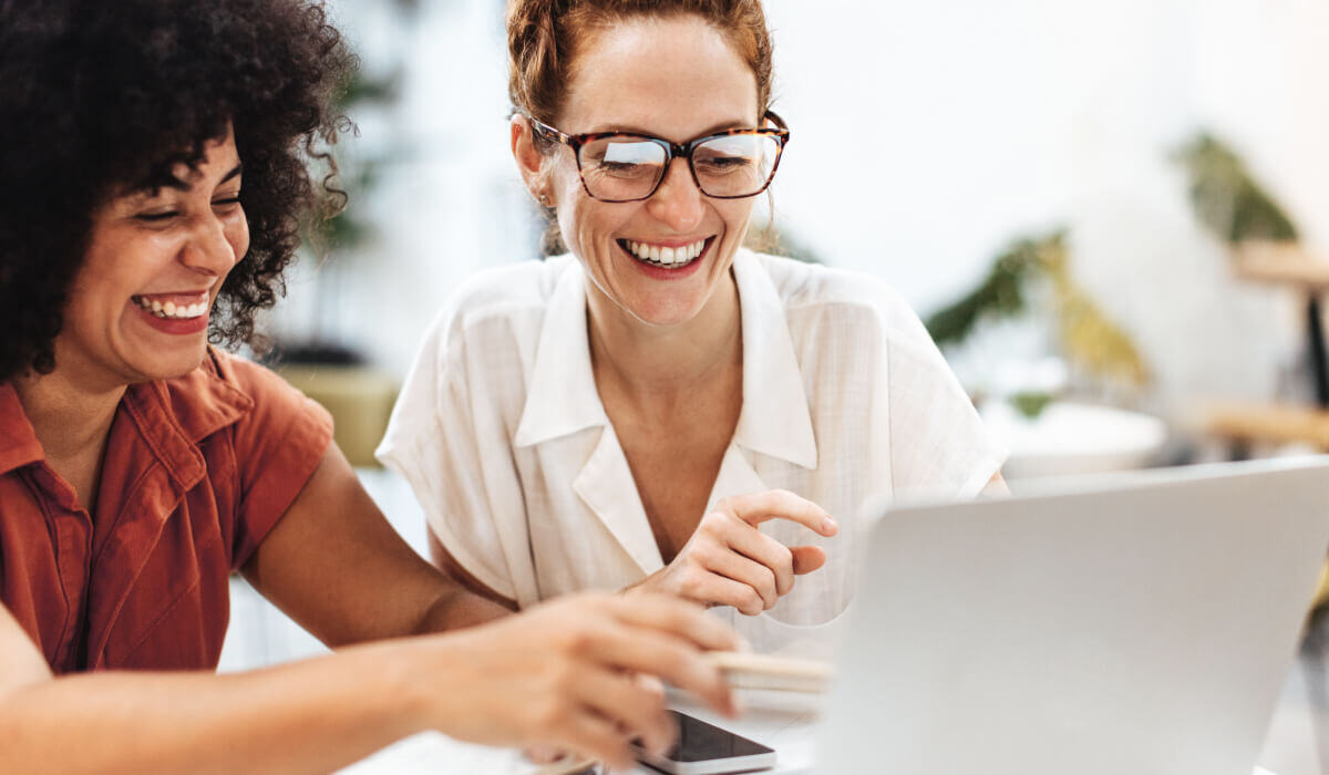 Two women, one with curly hair in a red blouse and the other wearing glasses, laugh joyfully while working together on a laptop, symbolizing the collaborative spirit and client-focused service of Vaulted's HubSpot web design process.
