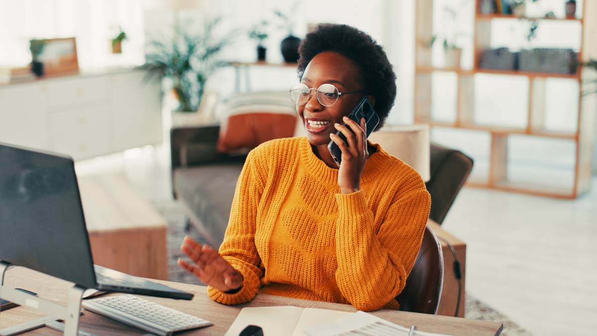 A smiling professional wearing a bright yellow sweater sits at a desk, talking on a smartphone and gesturing with one hand, while working on a laptop—representing the collaborative and connected experience possible with HubSpot’s Content Hub.