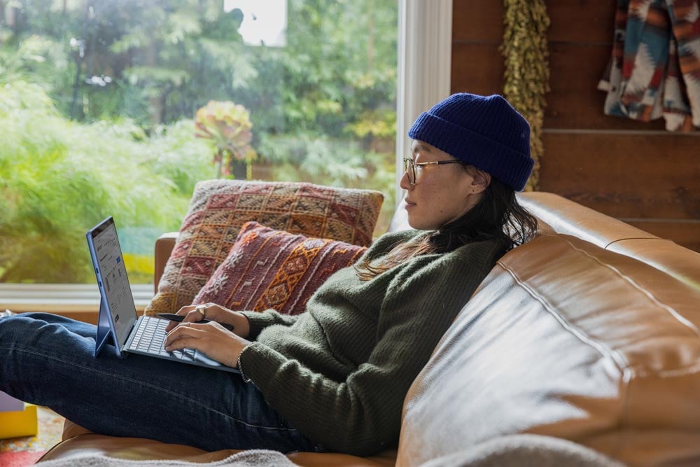 A person is sitting comfortably on a couch and focusing on their laptop screen, which shows their HubSpot account. Their fingers are actively typing, suggesting that they are managing or updating their account.