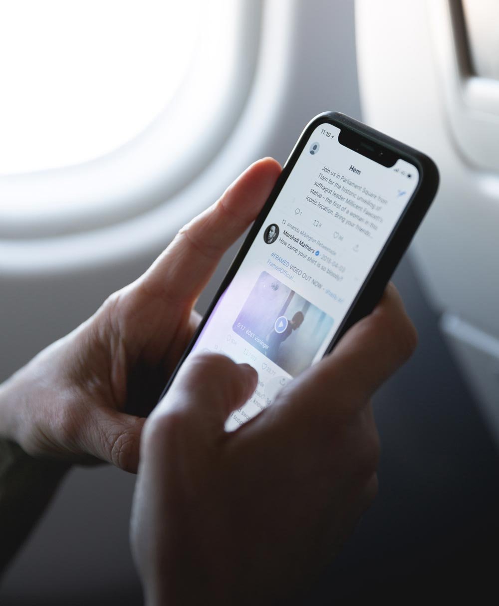 An individual on an airplane, cradling a smartphone in their hands. On the screen, vibrant icons of various social media platforms display recent updates, signifying their connection with the digital world even while in transit.