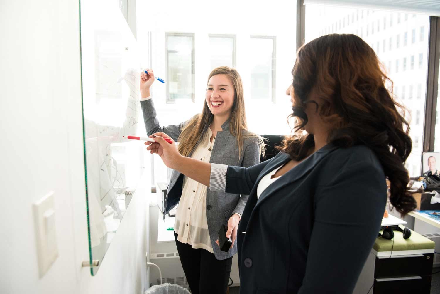 Two enthusiastic women collaborate on a whiteboard, crafting a dynamic HubSpot strategy for managing their distinct business units with a smile.